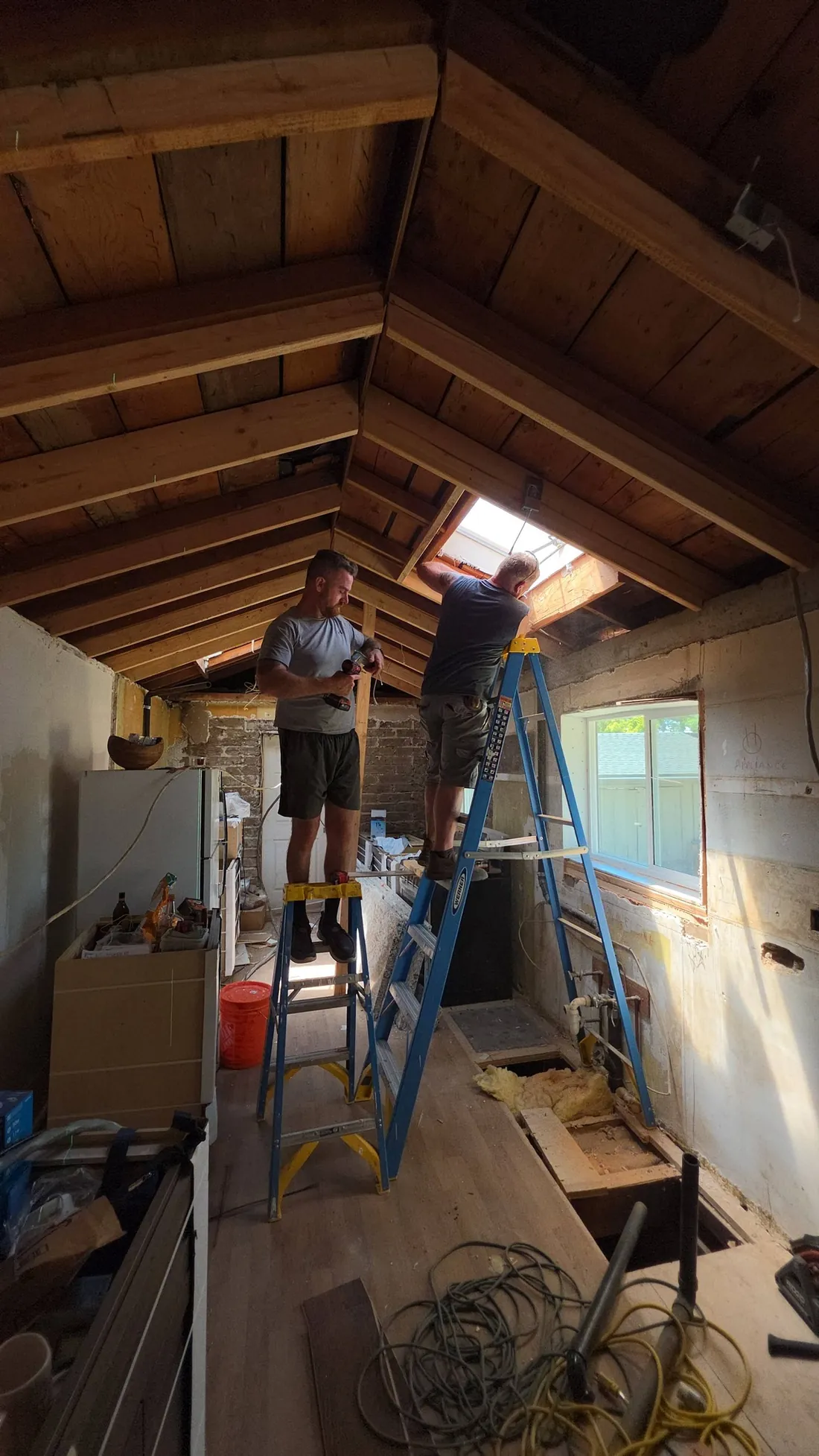 Two Pacific Builders craftsmen on ladders installing a skylight into a vaulted ceiling — natural light pouring through the new opening.